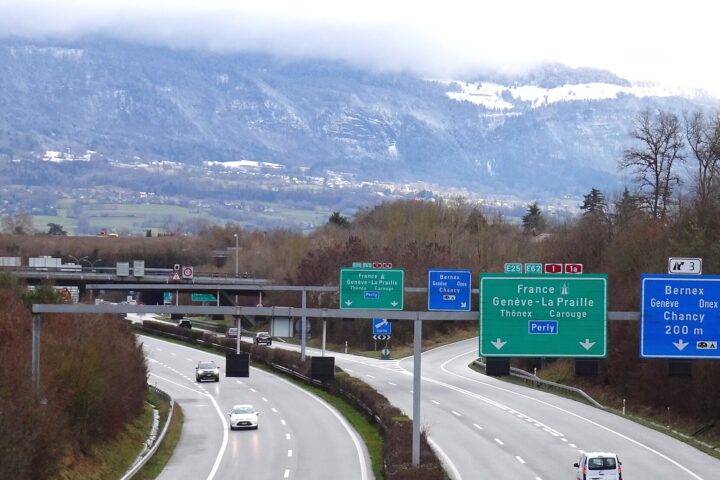 Cars approach the Bernex exit on Geneva's A1 motorway, with green direction signs and the snow-covered Salève behind.