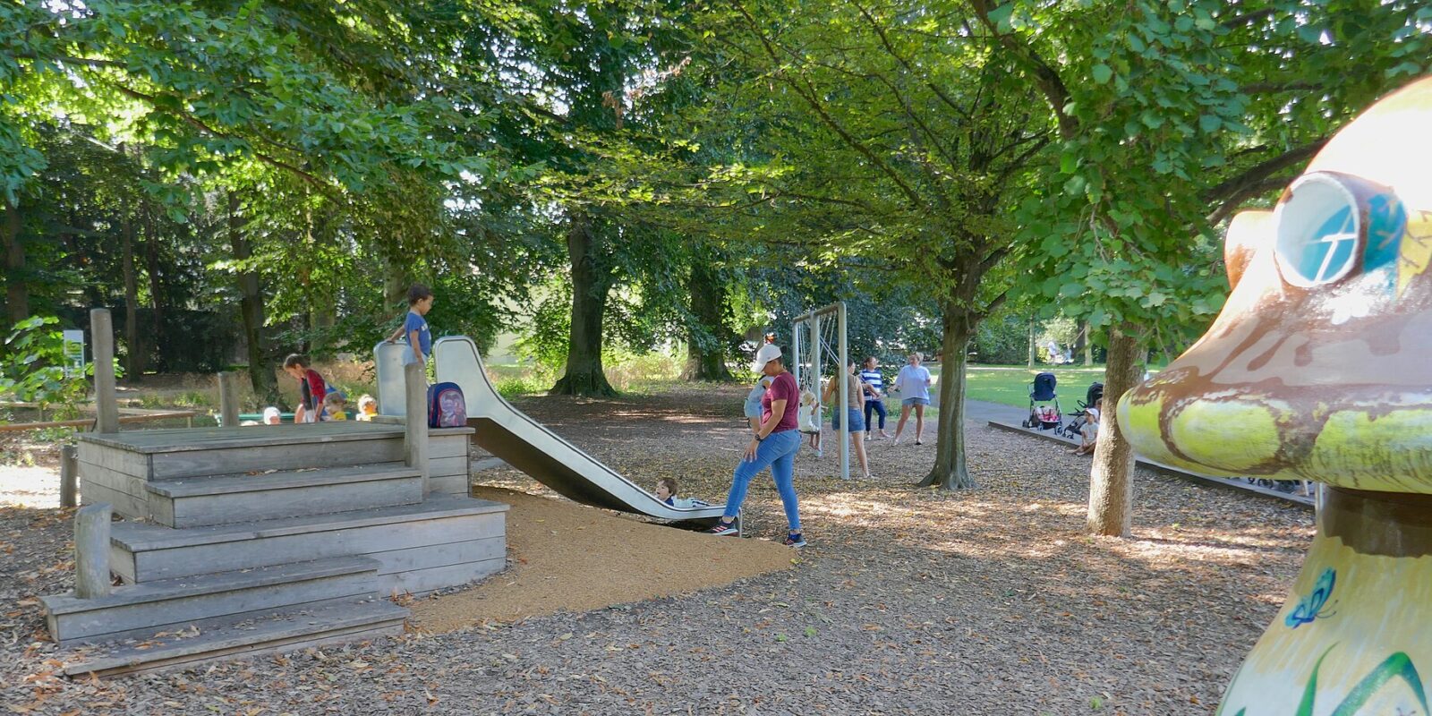 Children and adults gather around a slide beneath trees in Geneva's Parc La Grange, beside a colourful mushroom play structure.