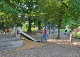 Children and adults gather around a slide beneath trees in Geneva's Parc La Grange, beside a colourful mushroom play structure.