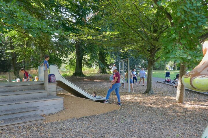 Children and adults gather around a slide beneath trees in Geneva's Parc La Grange, beside a colourful mushroom play structure.