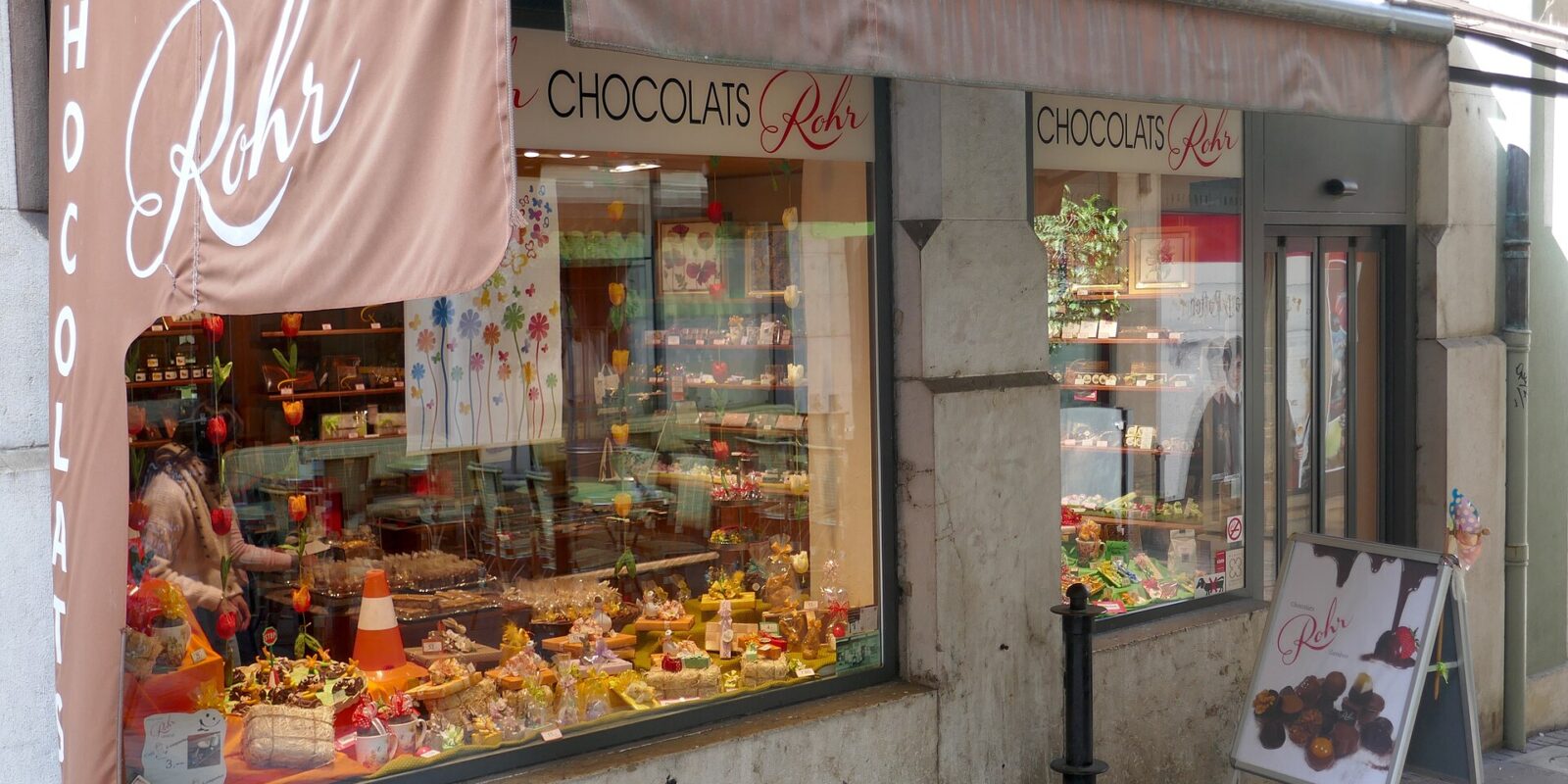Chocolats Rohr in Geneva displays chocolates and wrapped confections beneath a brown awning beside its glass entrance.