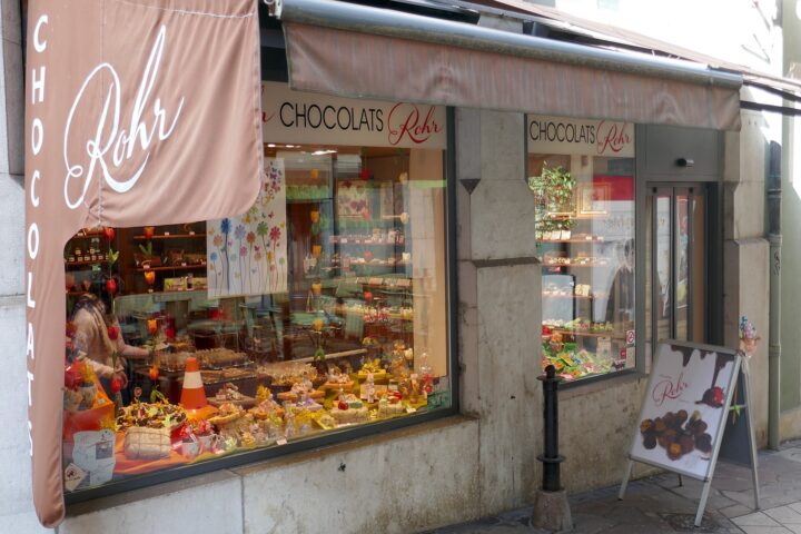 Chocolats Rohr in Geneva displays chocolates and wrapped confections beneath a brown awning beside its glass entrance.