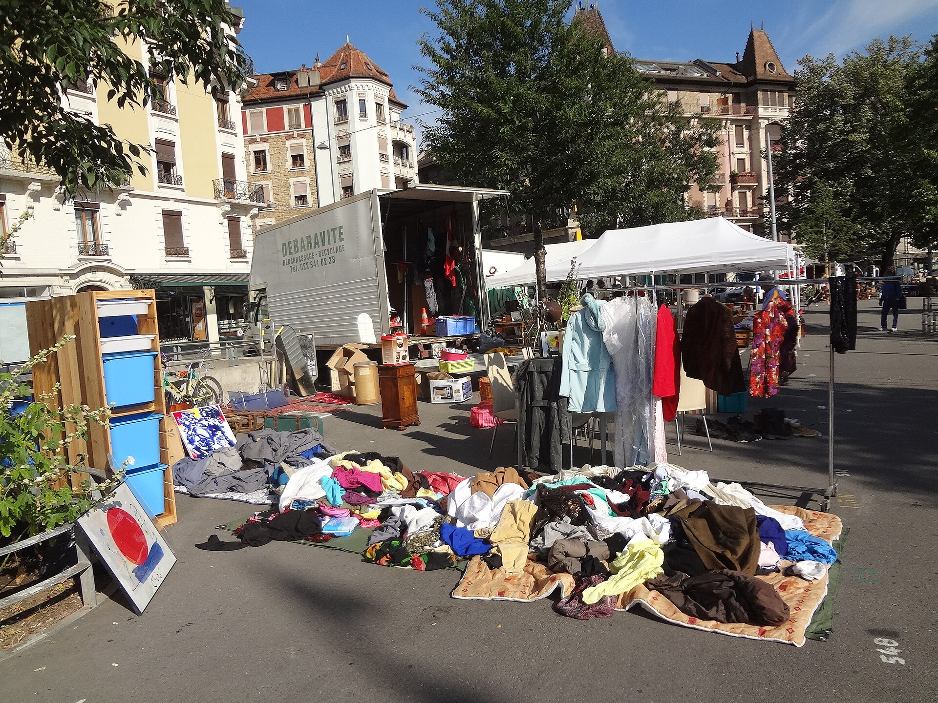 Clothes, furniture and household goods fill a flea-market stall beside trees and buildings at Plainpalais in Geneva.