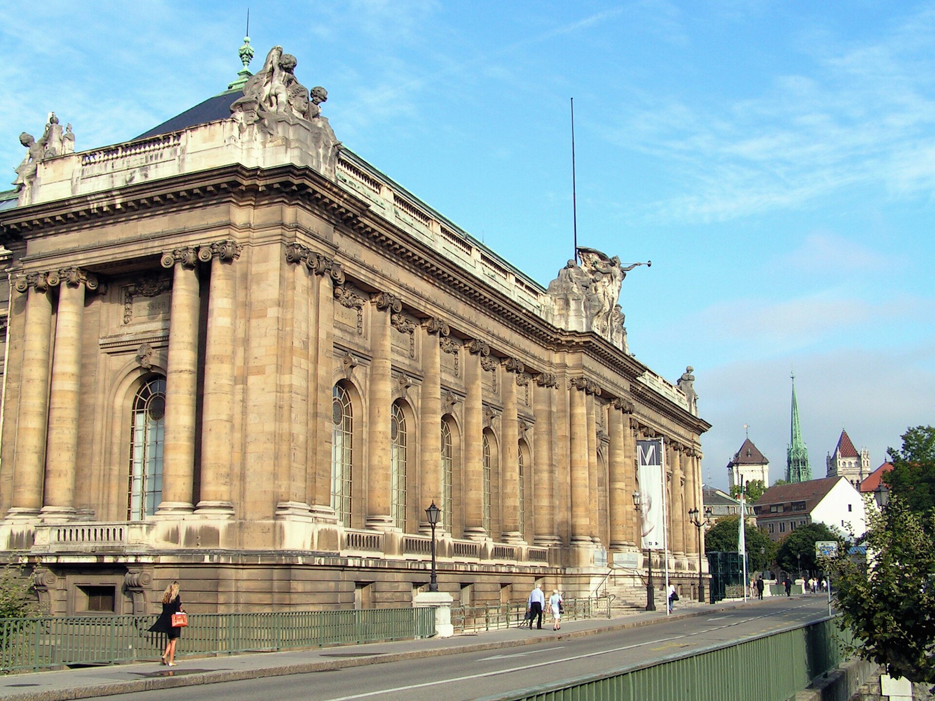 Columns, arched windows and rooftop sculptures line Geneva's Museum of Art and History, with cathedral towers visible beyond the street.