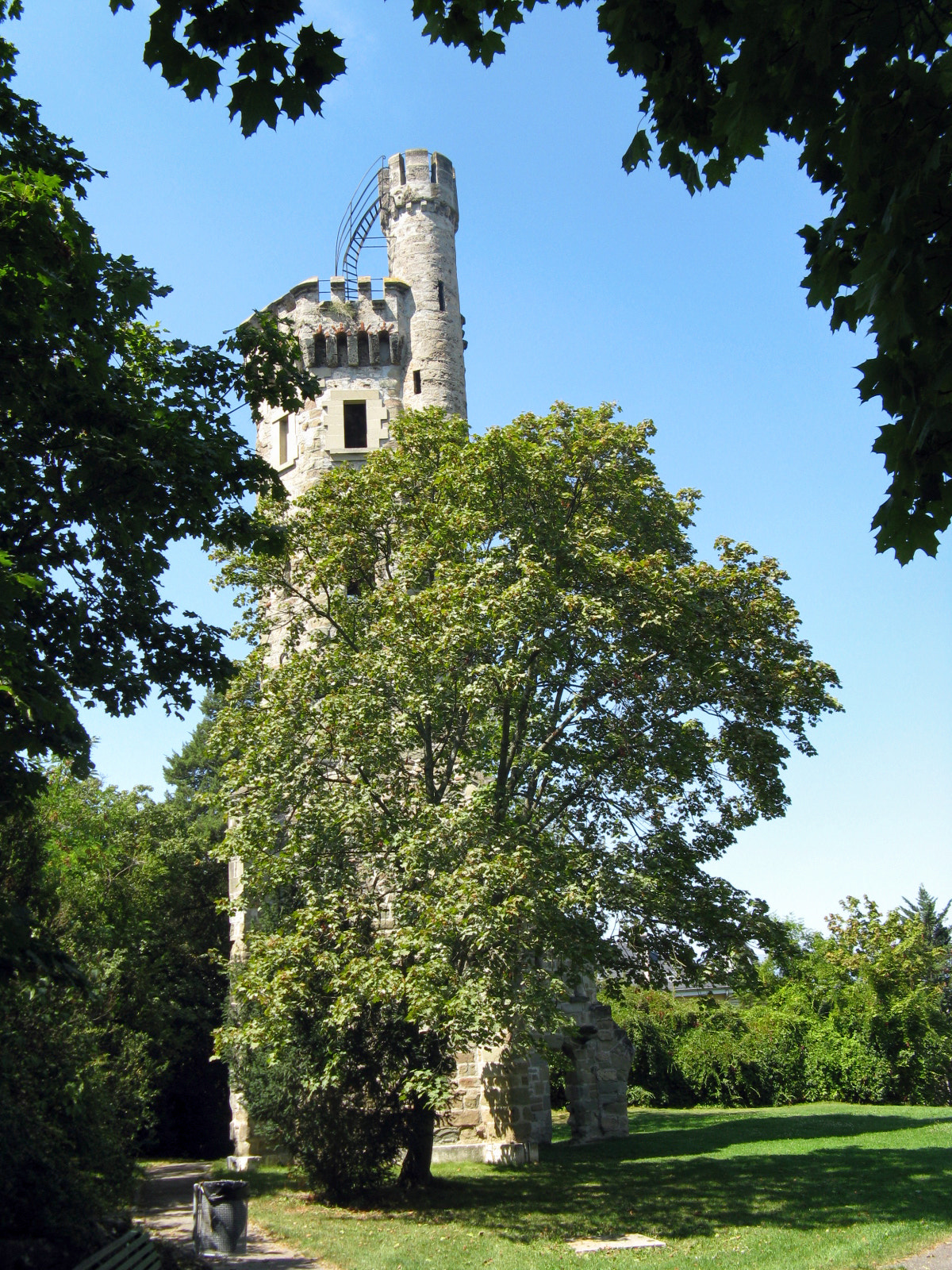 The crenellated Tour de Champel rises among leafy trees above a sunny lawn in Geneva, framed by overhanging branches.