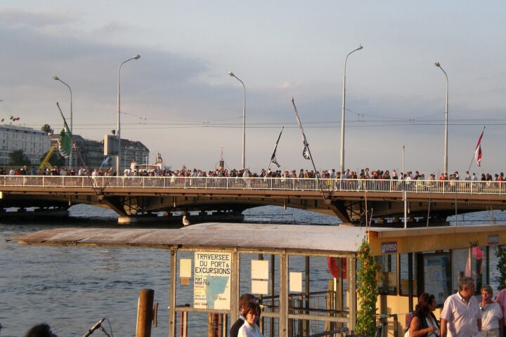 Crowds line Geneva's Pont du Mont-Blanc before the historic Fêtes fireworks, above a boat kiosk beside the water.