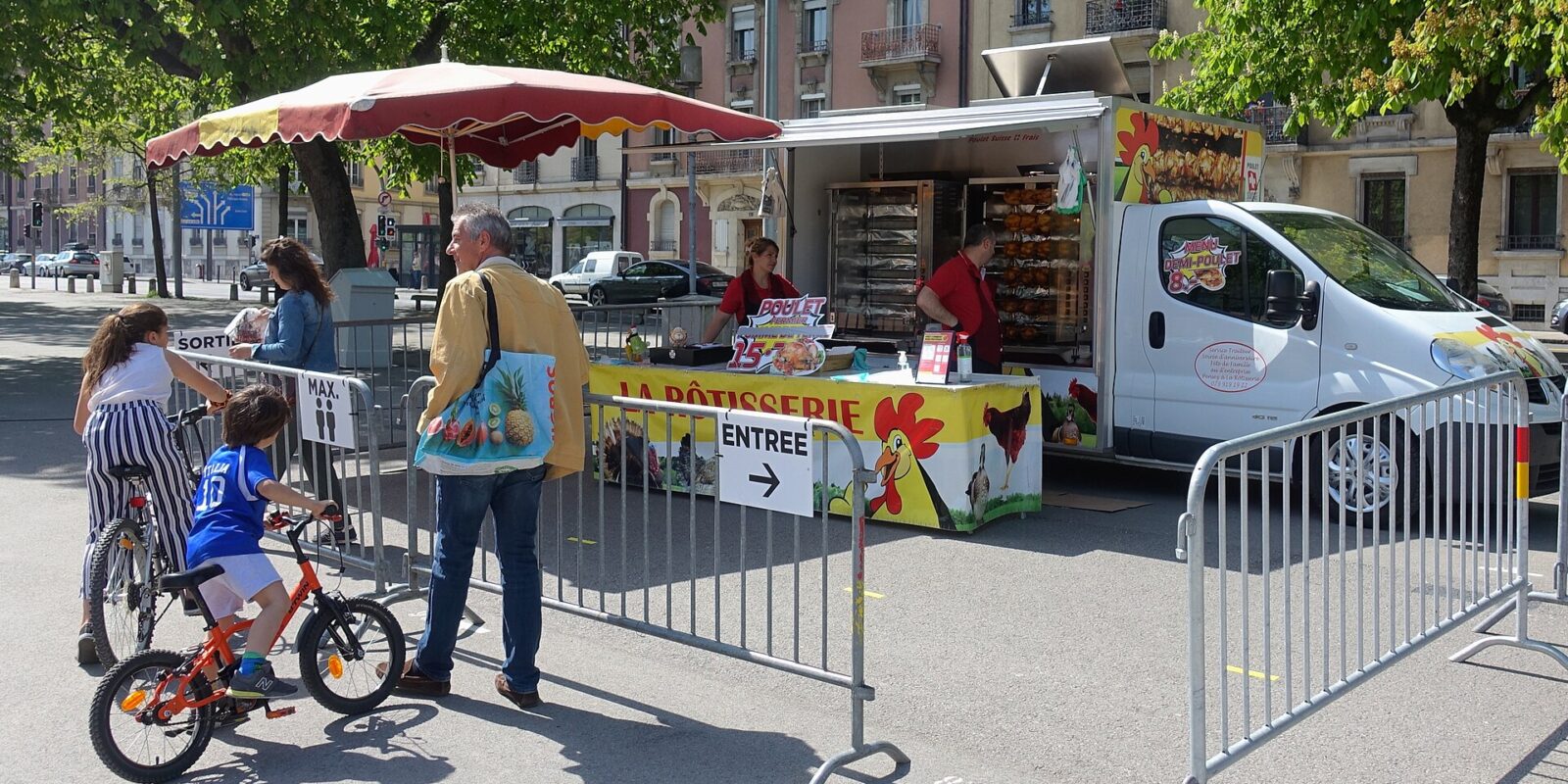 Customers wait by a rotisserie truck selling chicken at Geneva's Plainpalais market, with bicycles beside the queue barriers.