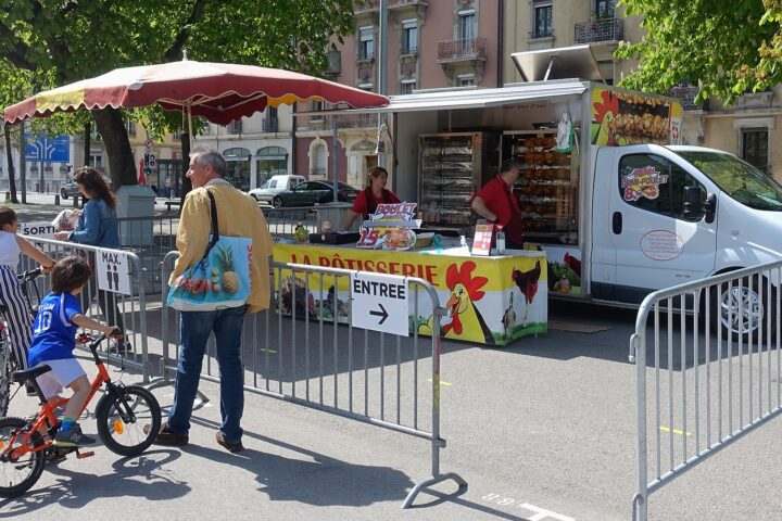 Customers wait by a rotisserie truck selling chicken at Geneva's Plainpalais market, with bicycles beside the queue barriers.