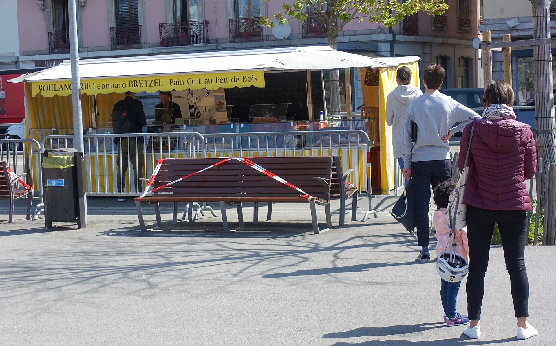 Customers wait beside a yellow-striped bakery stand at Geneva's Plainpalais market, with temporary barriers separating the counter from the pavement.