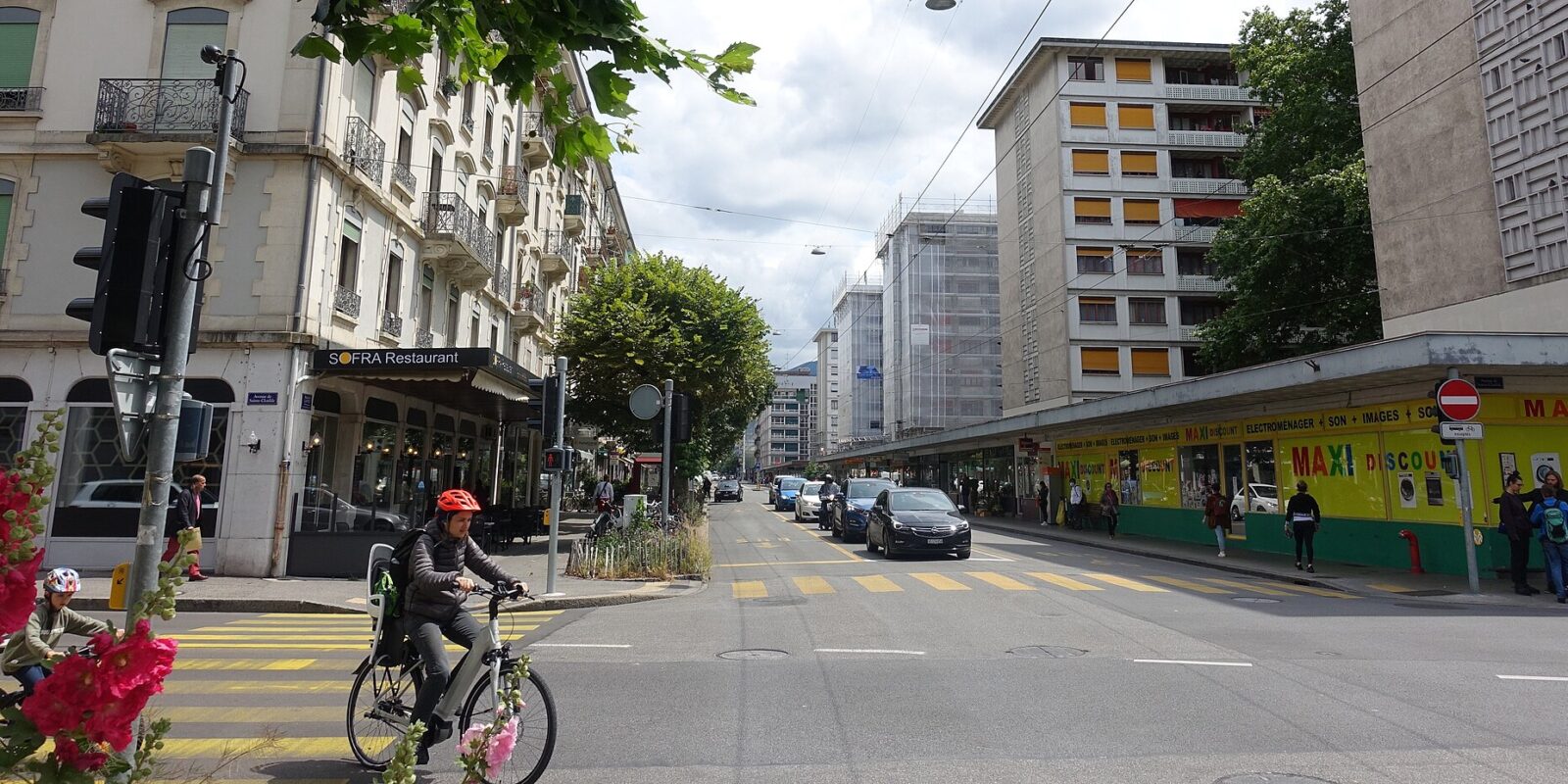A cyclist crosses Boulevard Carl-Vogt in Geneva, with a corner restaurant and discount shops lining the street.