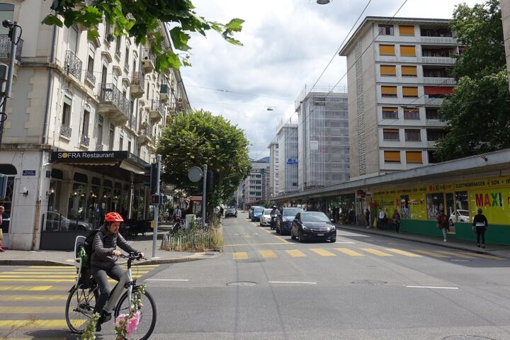 A cyclist crosses Boulevard Carl-Vogt in Geneva, with a corner restaurant and discount shops lining the street.