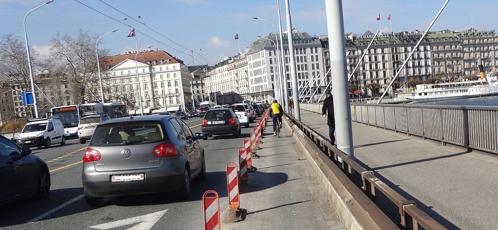 A cyclist rides across Geneva's Pont du Mont-Blanc beside traffic, with waterfront hotels and a white paddle steamer beyond the railing.
