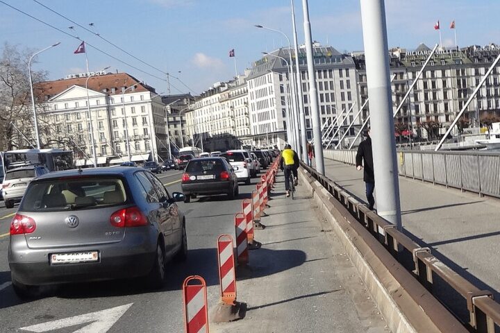 A cyclist rides across Geneva's Pont du Mont-Blanc beside traffic, with waterfront hotels and a white paddle steamer beyond the railing.
