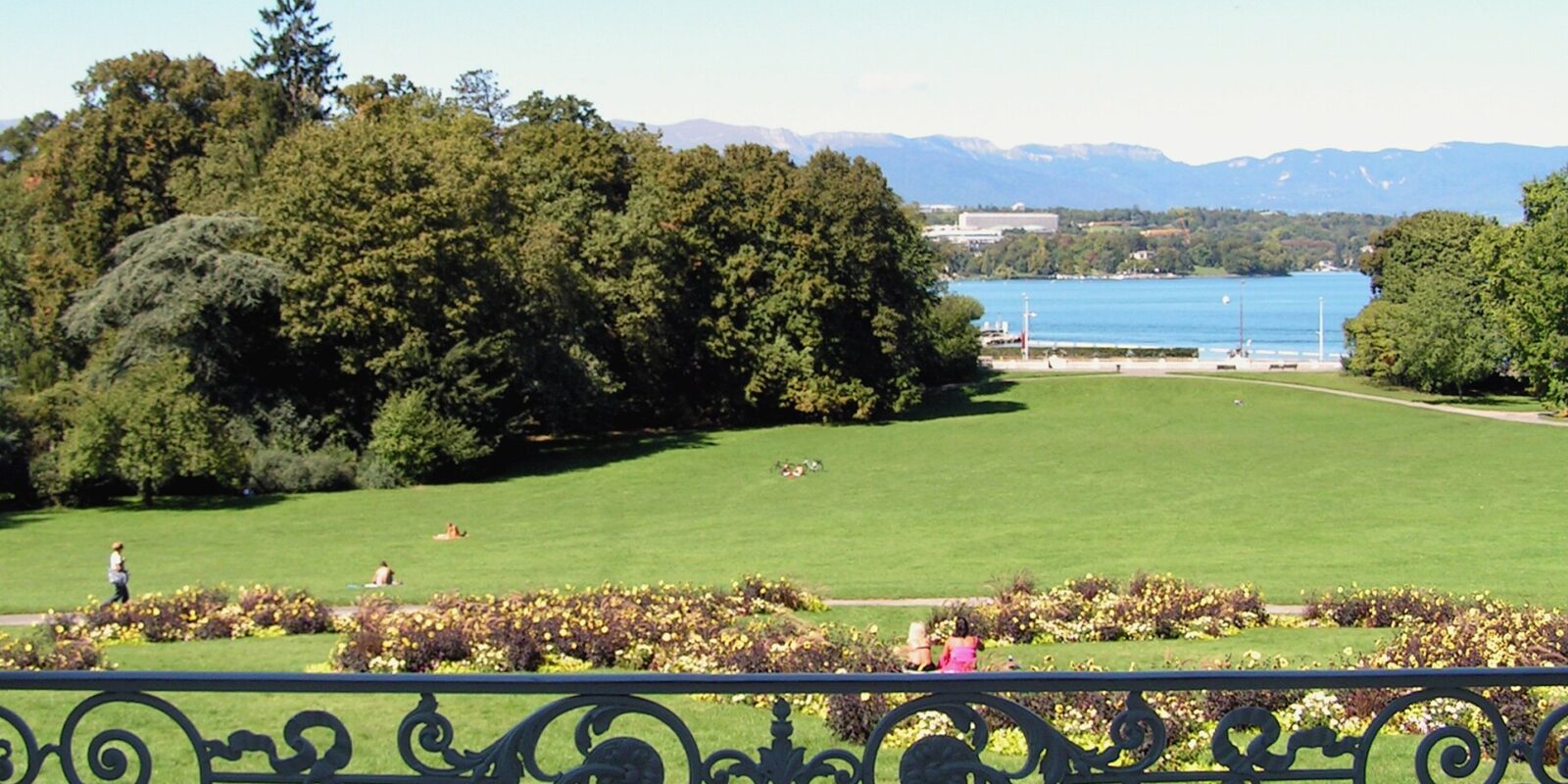 A decorative terrace railing frames flower beds and sunny lawns descending toward Lake Geneva from Parc La Grange in September.