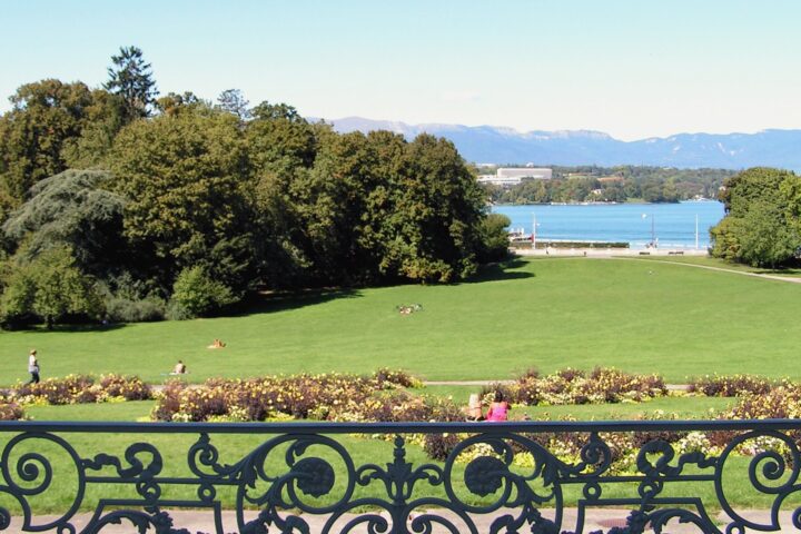 A decorative terrace railing frames flower beds and sunny lawns descending toward Lake Geneva from Parc La Grange in September.
