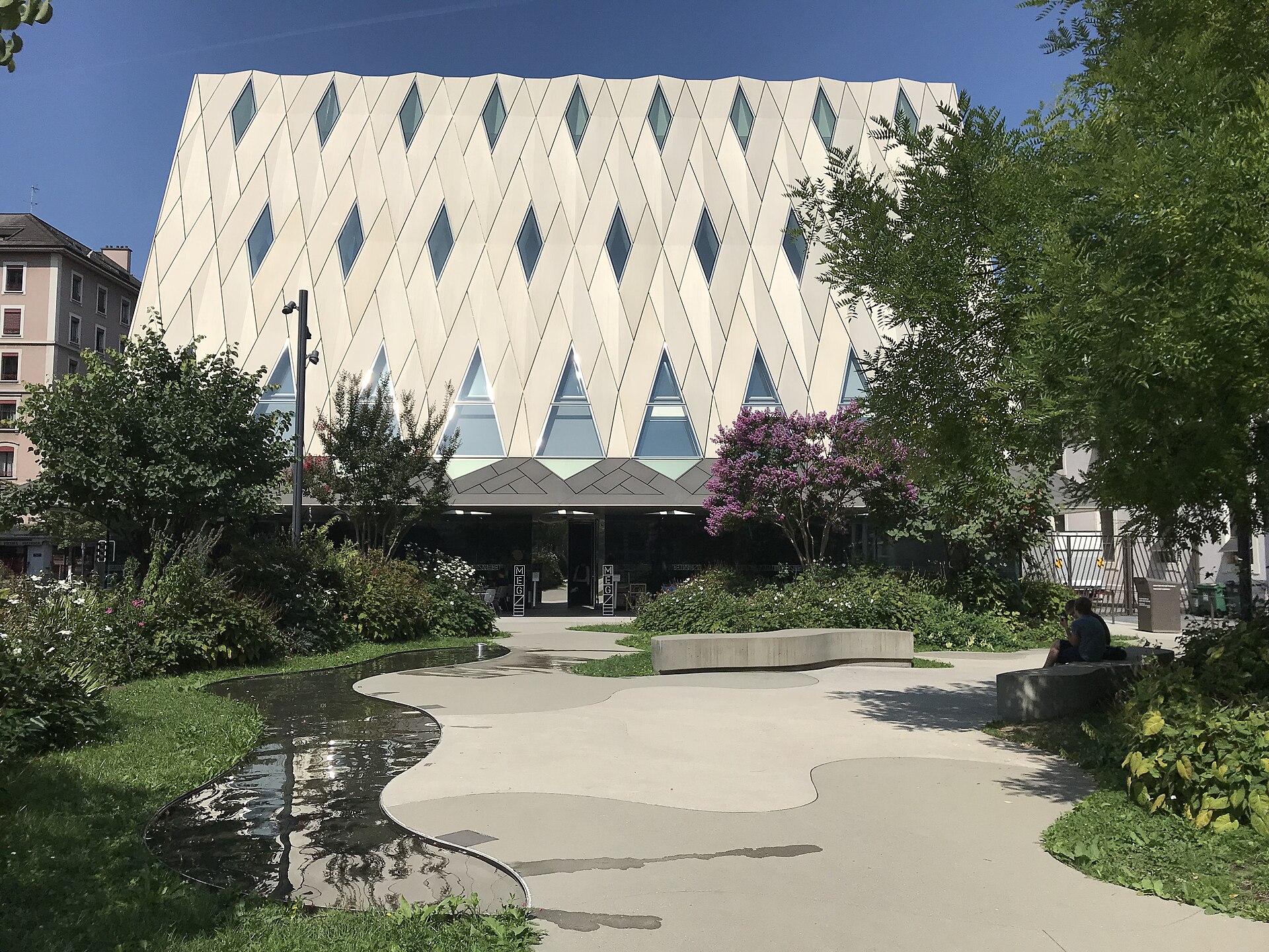 Diamond-patterned white facade of the Musée d'ethnographie de Genève behind a landscaped garden with a wide paved path leading toward the glazed ground floor.
