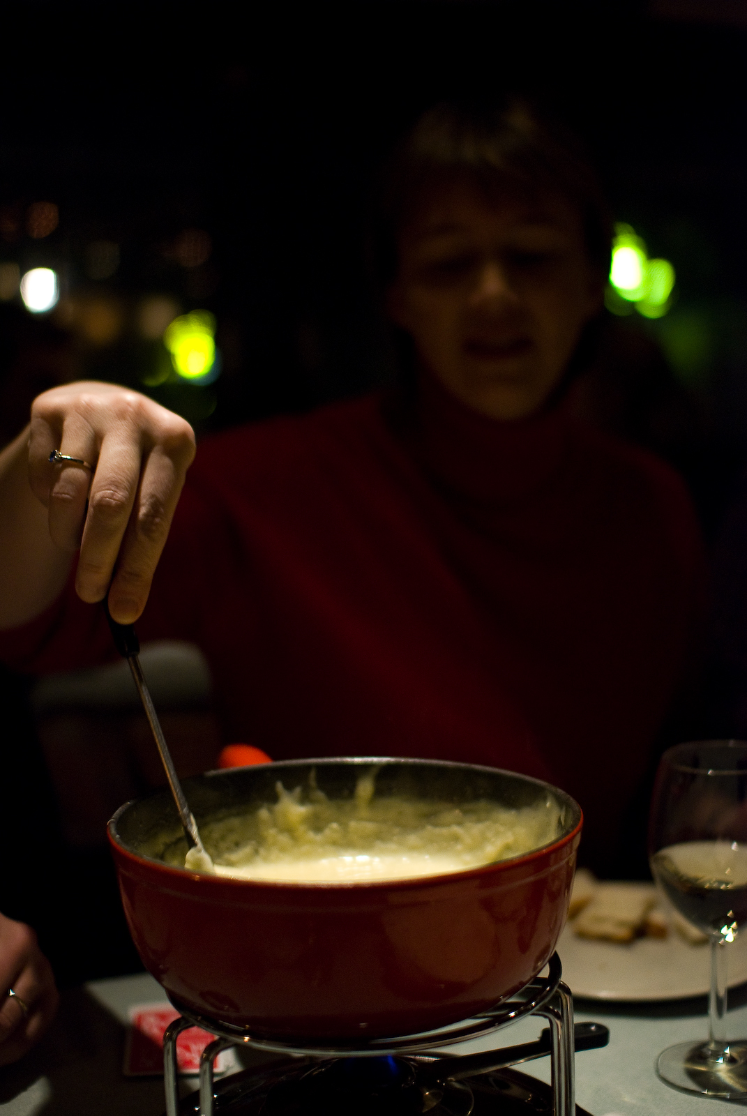 A diner dips a long fork into cheese fondue in a red caquelon kept warm over a small blue flame.