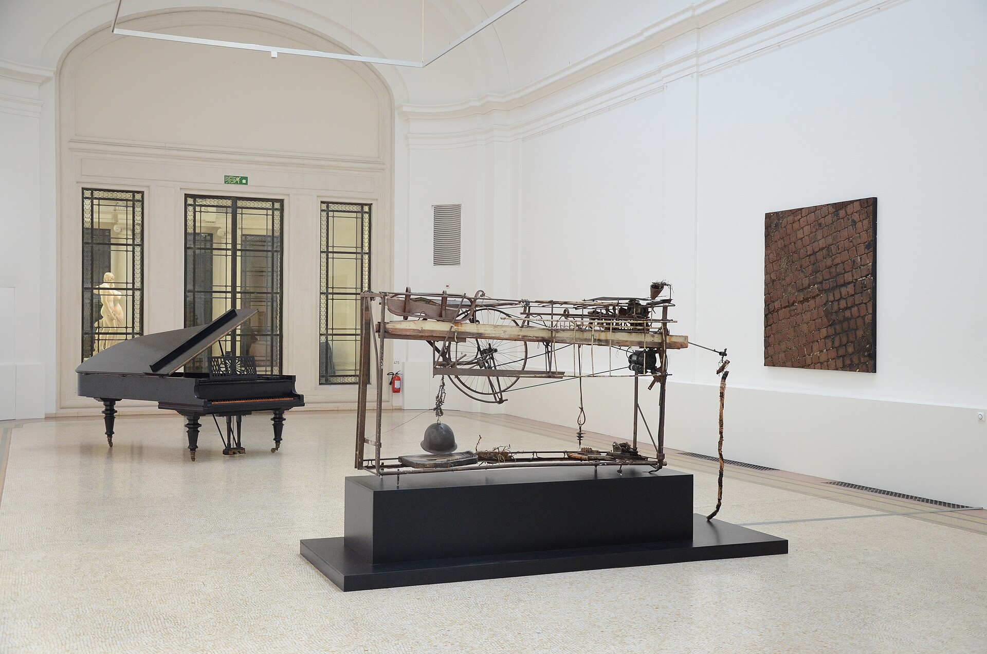 An exposed mechanical structure, a grand piano and a textured wall work occupy a bright gallery inside Geneva’s MAH.