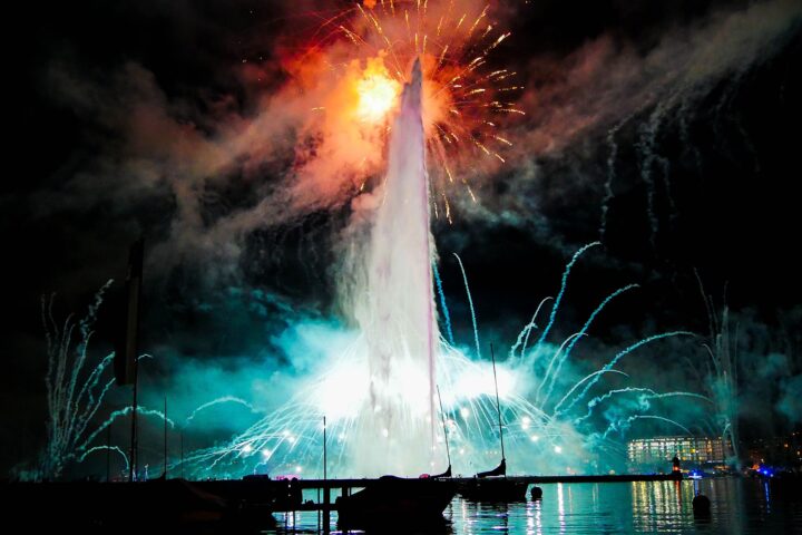 Fireworks burst above Geneva’s illuminated Jet d’Eau, with moored sailboats silhouetted against coloured reflections on the harbour.