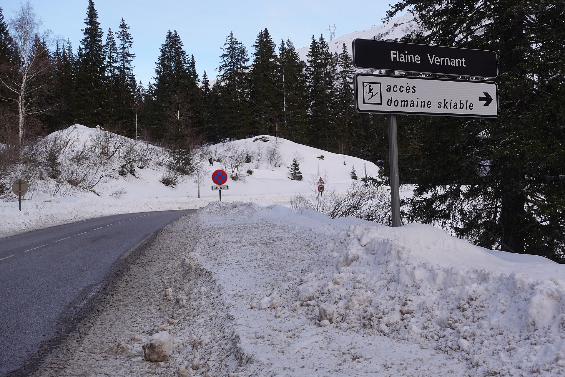 A Flaine Vernant sign points toward the ski area beside a snowy mountain road lined with evergreen trees.