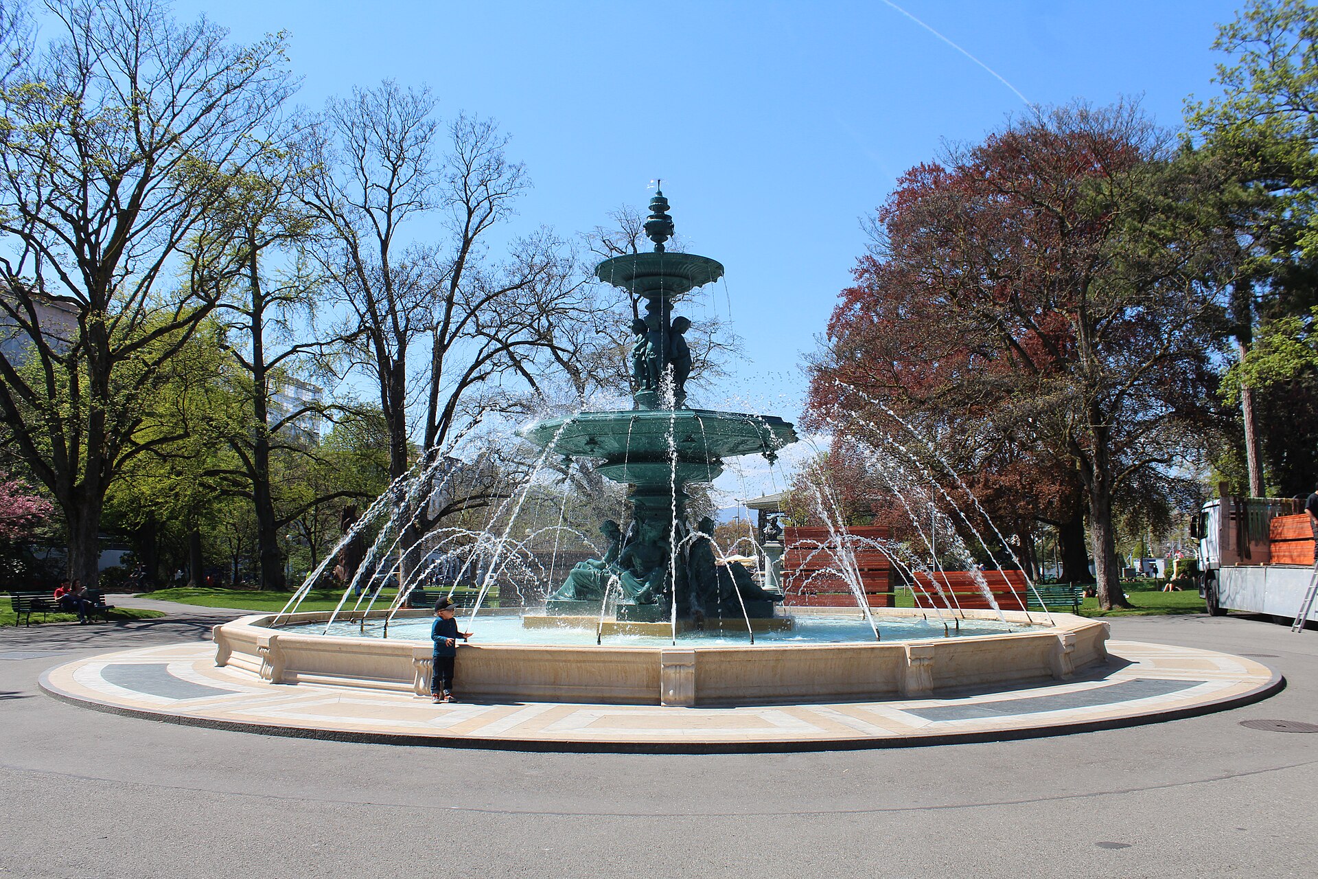 An ornate dark green fountain spraying water in the middle of the Jardin Anglais in Geneva, among spring trees