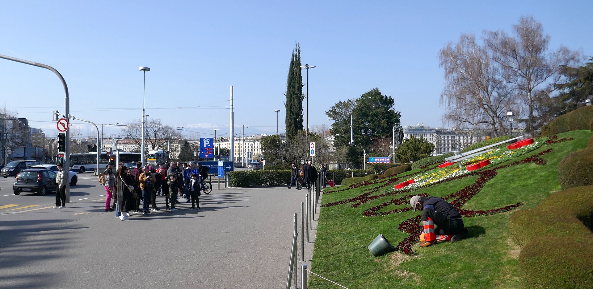 A city gardener kneeling to tend the planting of the Flower Clock in Geneva while a group of visitors walks past on the quay