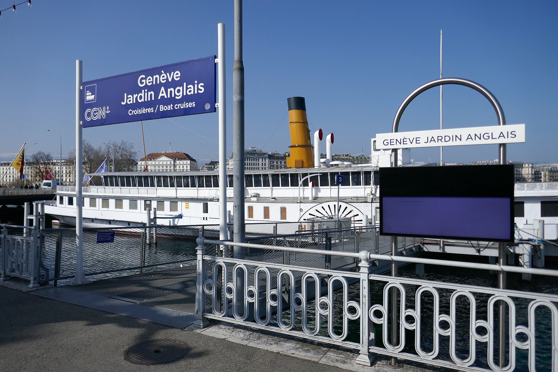 The Genève Jardin Anglais boat landing stage with its blue sign, and a white paddle steamer with a yellow funnel moored alongside