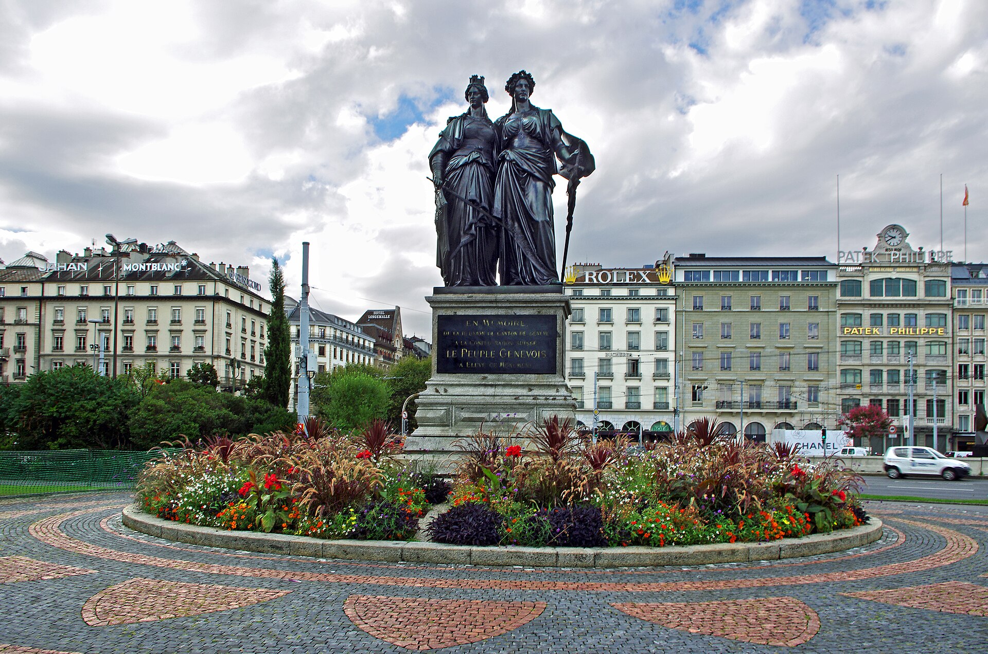 The Monument national in Geneva, two bronze female figures on a stone pedestal above a circular bed of red flowers, with lakefront buildings behind