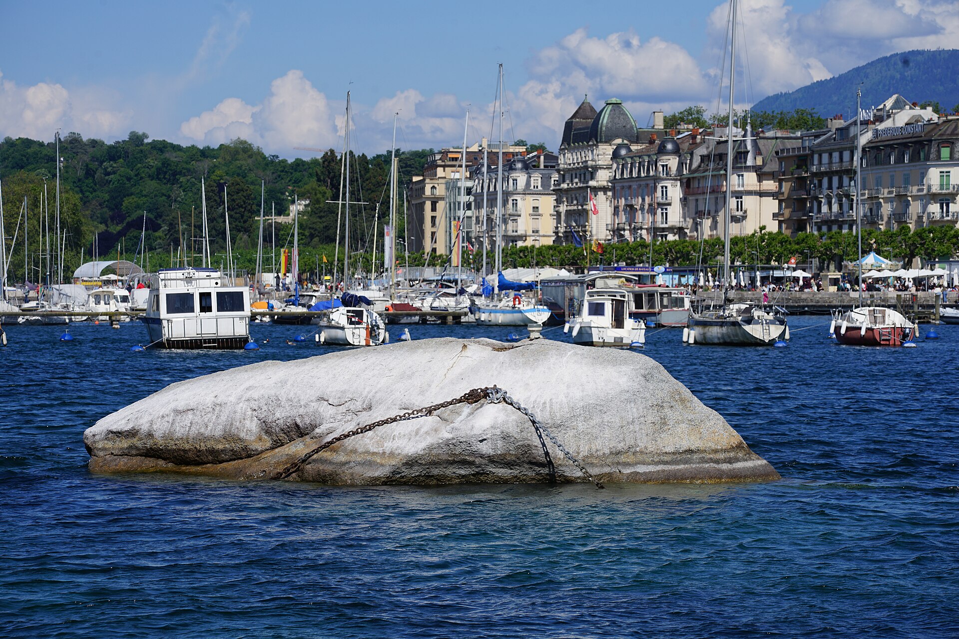 The large pale granite Pierre du Niton boulder rising from the blue water of Geneva's harbour, with moored boats and lakefront buildings behind