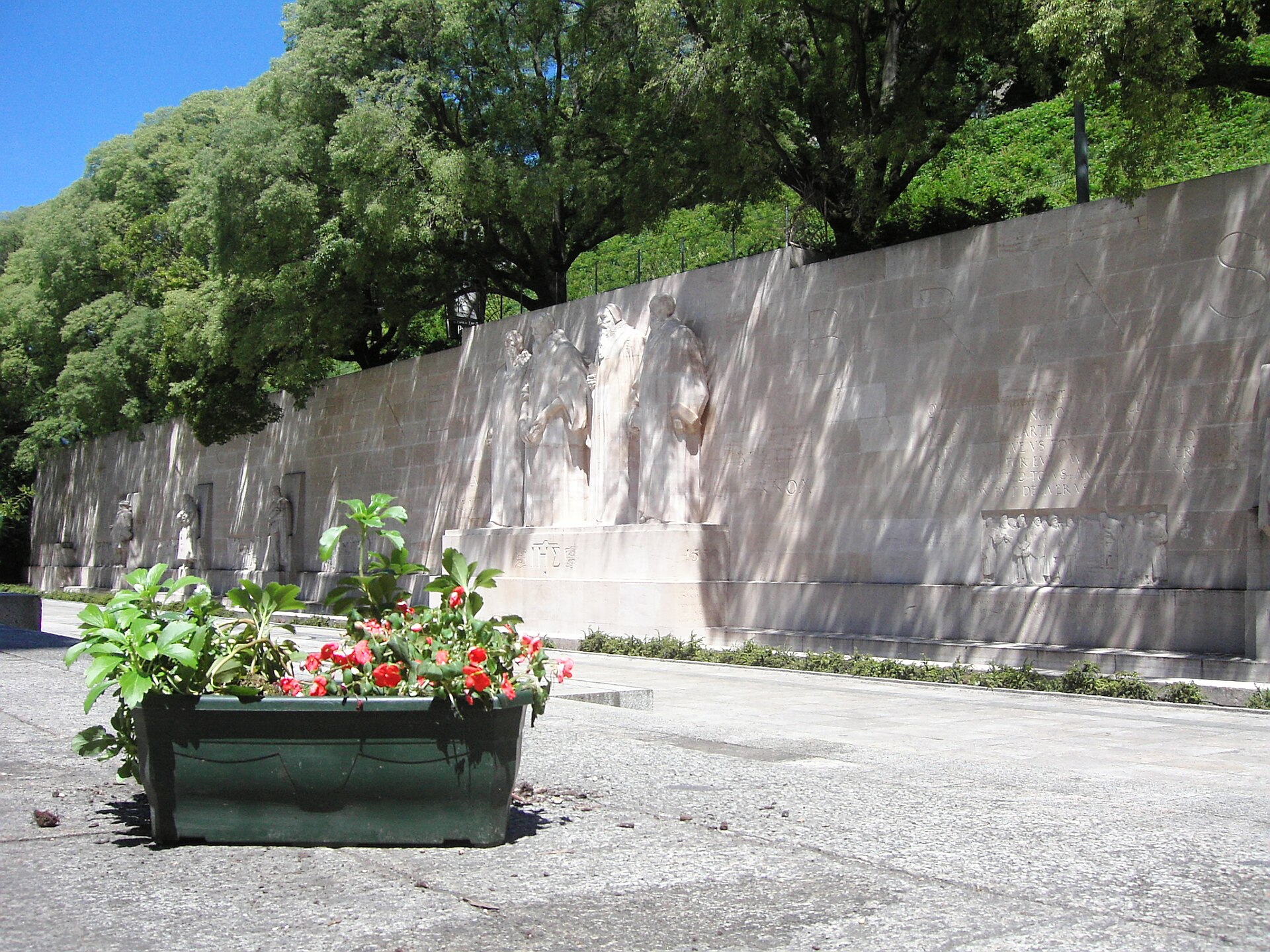 A flower planter stands before the Reformation Wall and its four central statues beneath leafy trees in Geneva.