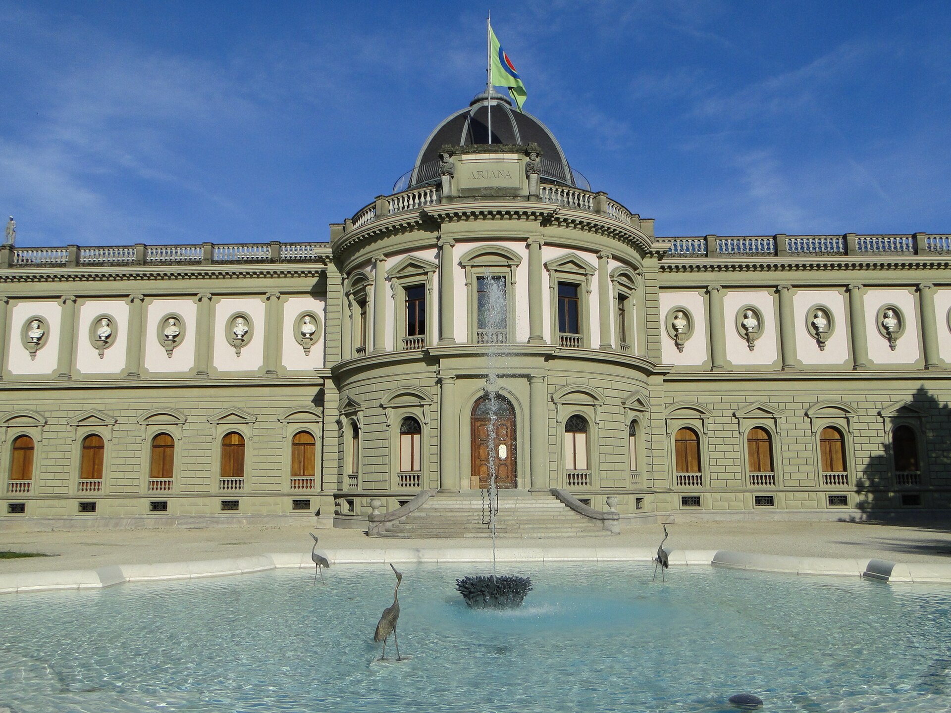 A fountain and bird sculptures stand before the domed Ariana museum in Geneva under a clear blue sky.