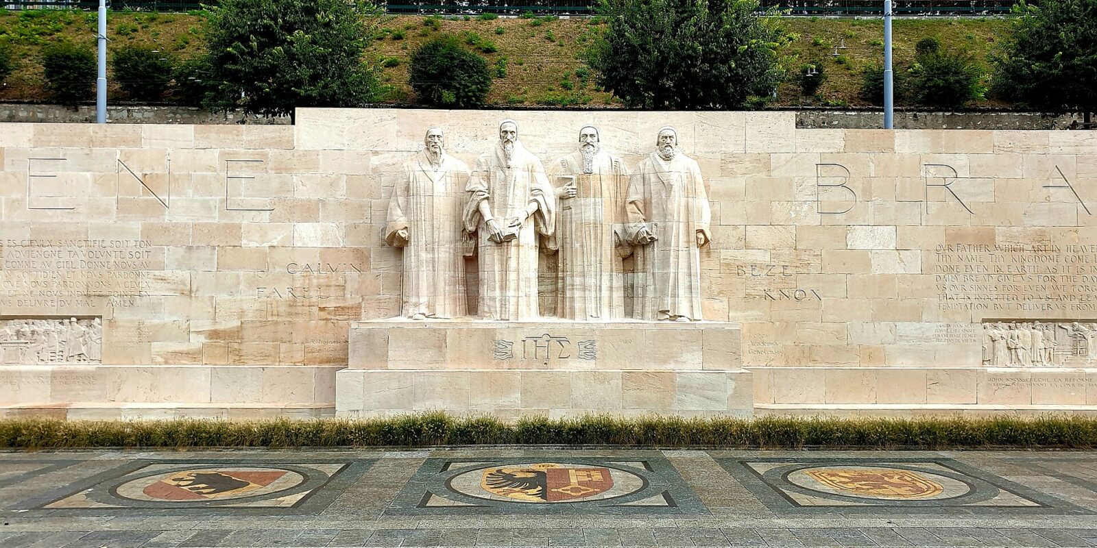 Four stone reformers stand above heraldic pavement mosaics at Geneva's Reformation Wall, with inscriptions stretching across the pale stone.