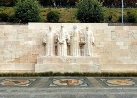 Four stone reformers stand above heraldic pavement mosaics at Geneva's Reformation Wall, with inscriptions stretching across the pale stone.