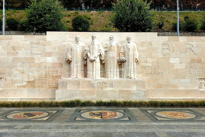 Four stone reformers stand above heraldic pavement mosaics at Geneva's Reformation Wall, with inscriptions stretching across the pale stone.