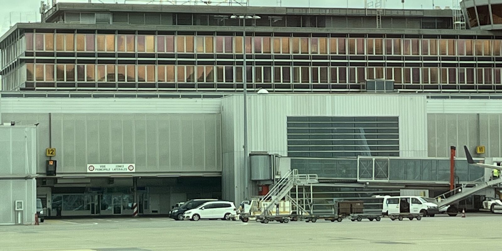 Geneva Airport's terminal building seen from the apron, with the rooftop GENEVE AEROPORT lettering, control tower, jet bridges and ground vehicles under an overcast sky.