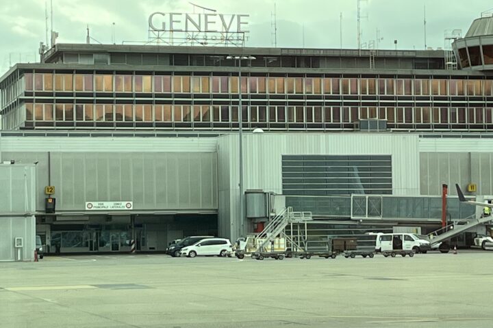 Geneva Airport's terminal building seen from the apron, with the rooftop GENEVE AEROPORT lettering, control tower, jet bridges and ground vehicles under an overcast sky.
