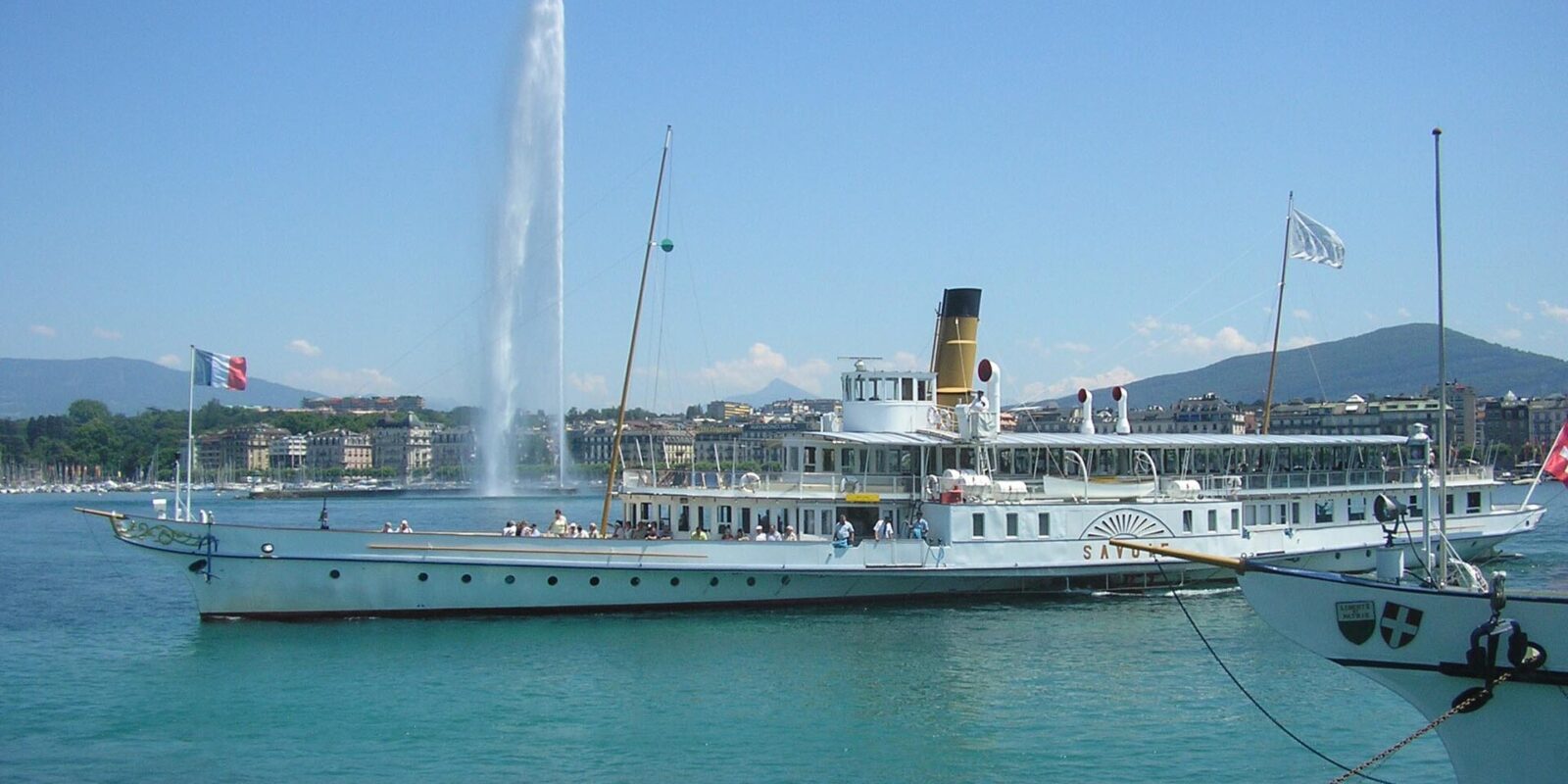 The paddle steamer Savoie moored in Geneva's harbour on a sunny day, with the Jet d'Eau rising behind it and the far shore beyond