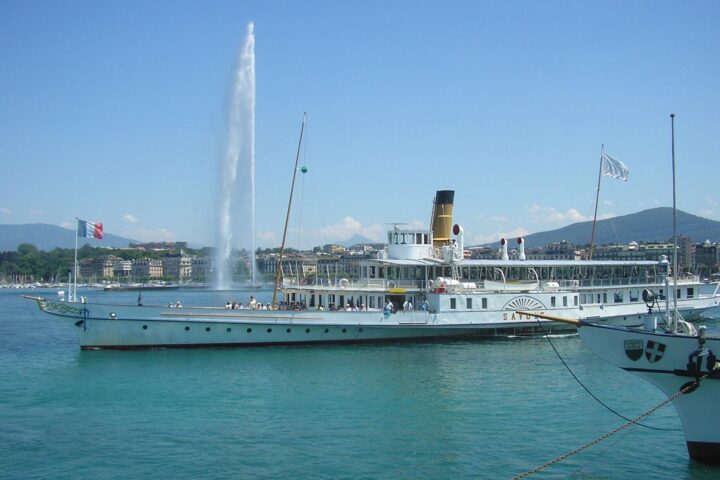 The paddle steamer Savoie moored in Geneva's harbour on a sunny day, with the Jet d'Eau rising behind it and the far shore beyond