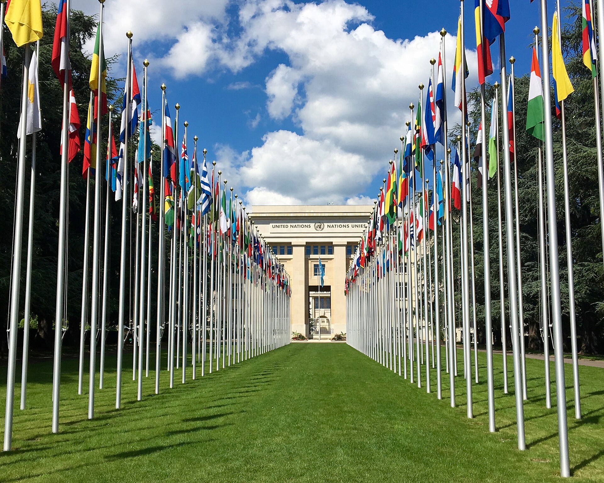 The avenue of national flags on tall poles leading to the Palais des Nations in Geneva, under a blue sky with clouds