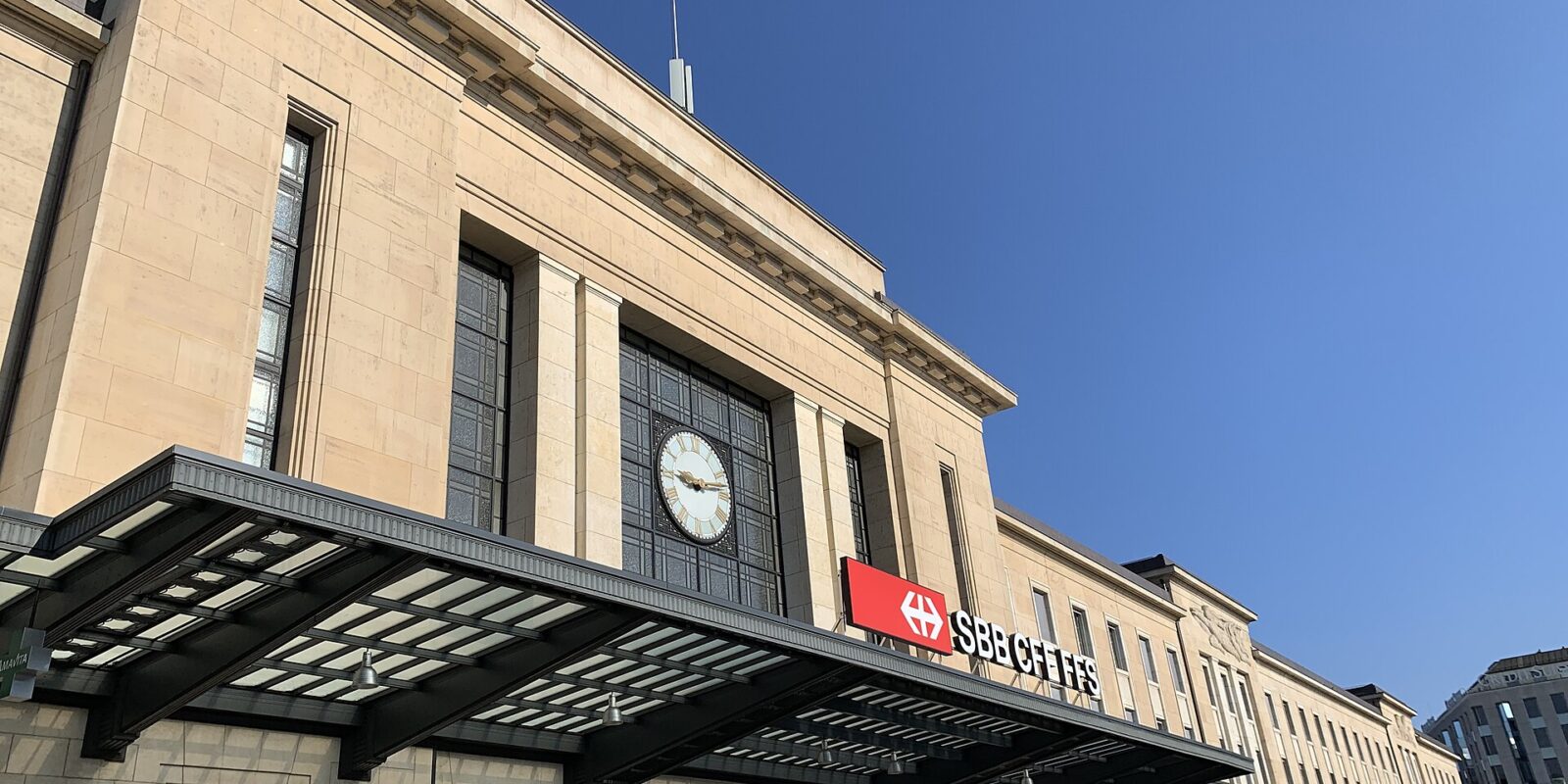 Geneva Cornavin station's clock and SBB sign rise above a broad glass entrance canopy under a clear blue sky.