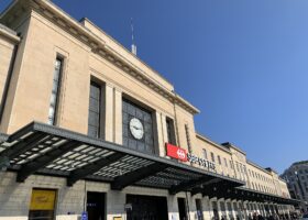 Geneva Cornavin station's clock and SBB sign rise above a broad glass entrance canopy under a clear blue sky.