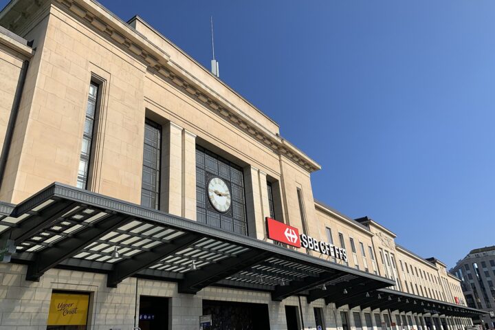 Geneva Cornavin station's clock and SBB sign rise above a broad glass entrance canopy under a clear blue sky.
