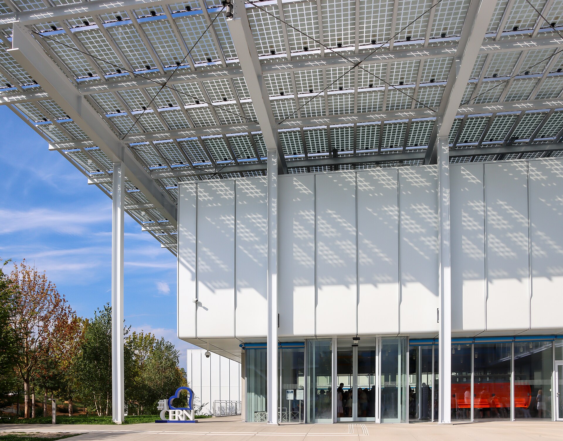 The white CERN Science Gateway pavilion in Meyrin under its large canopy of solar panels, with trees on the left