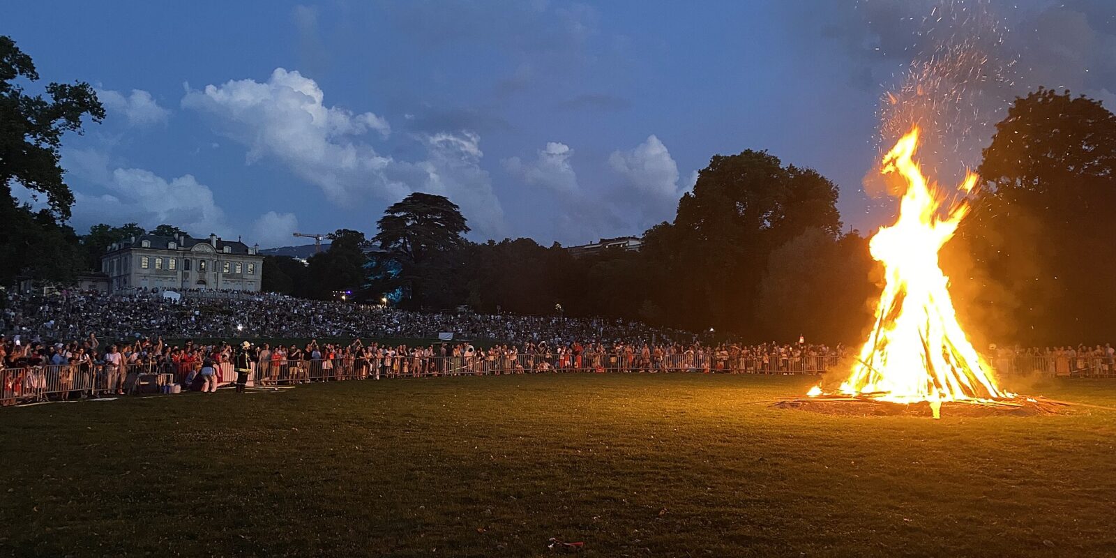 A large bonfire burning on the lawn of Parc La Grange in Geneva on 1 August 2024, with a crowd watching and the Villa La Grange behind at dusk