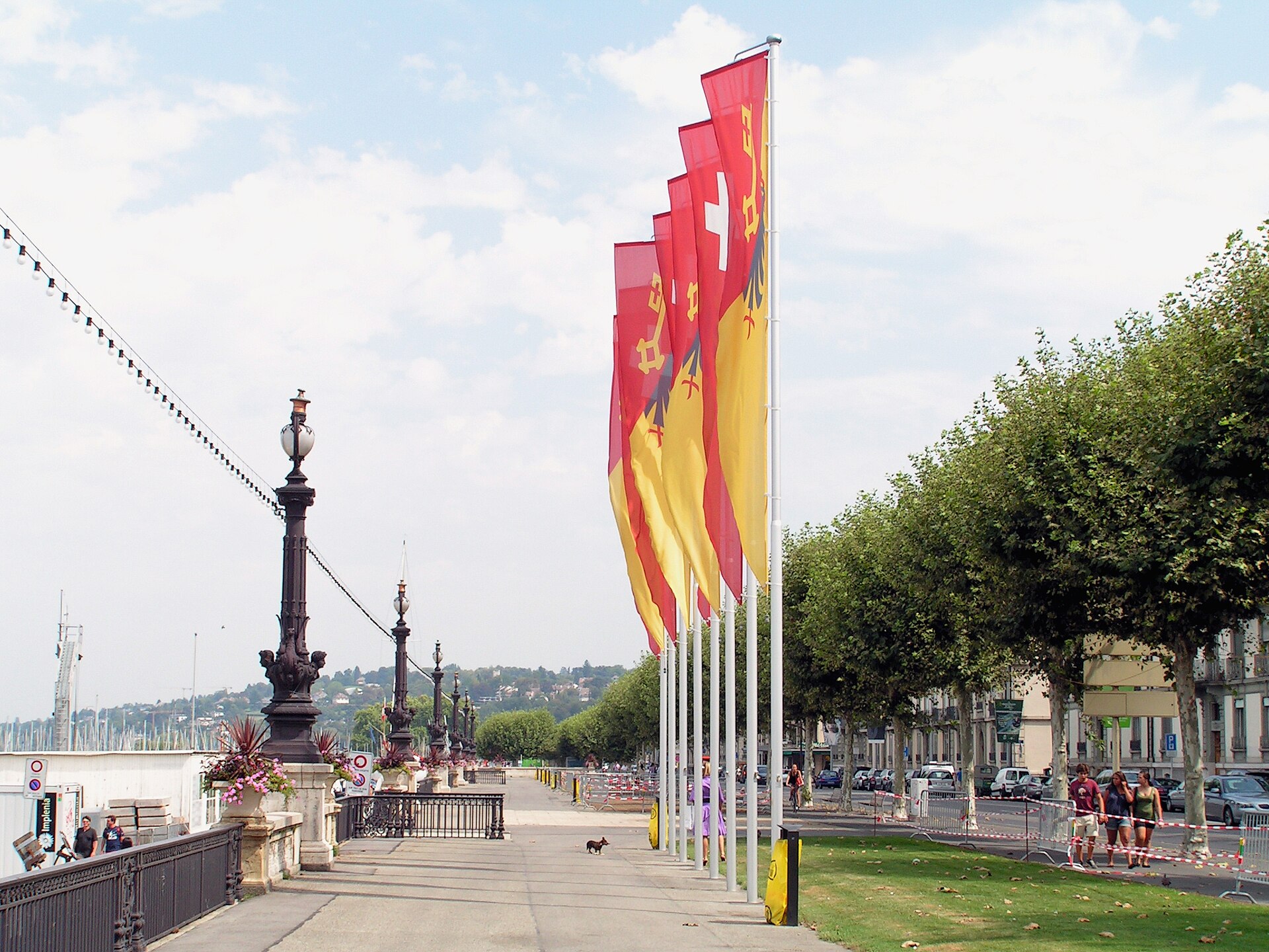 A row of red and yellow Geneva and Swiss flags along the wide, quiet lakeside promenade of the Quai Gustave-Ador, with plane trees and lamp posts