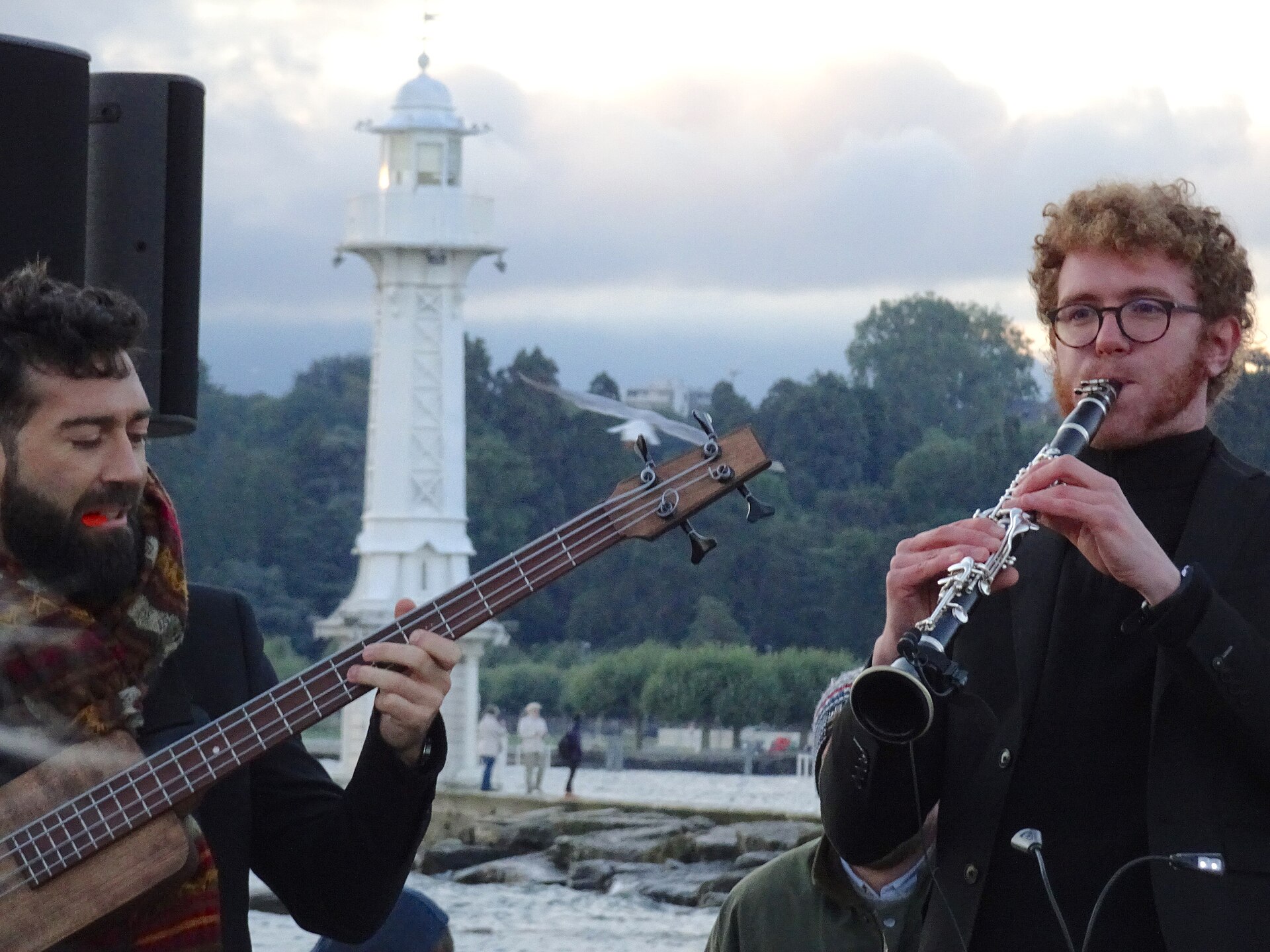 A bass player and a clarinettist performing on the jetty of the Bains des Pâquis, with the white Pâquis lighthouse behind them