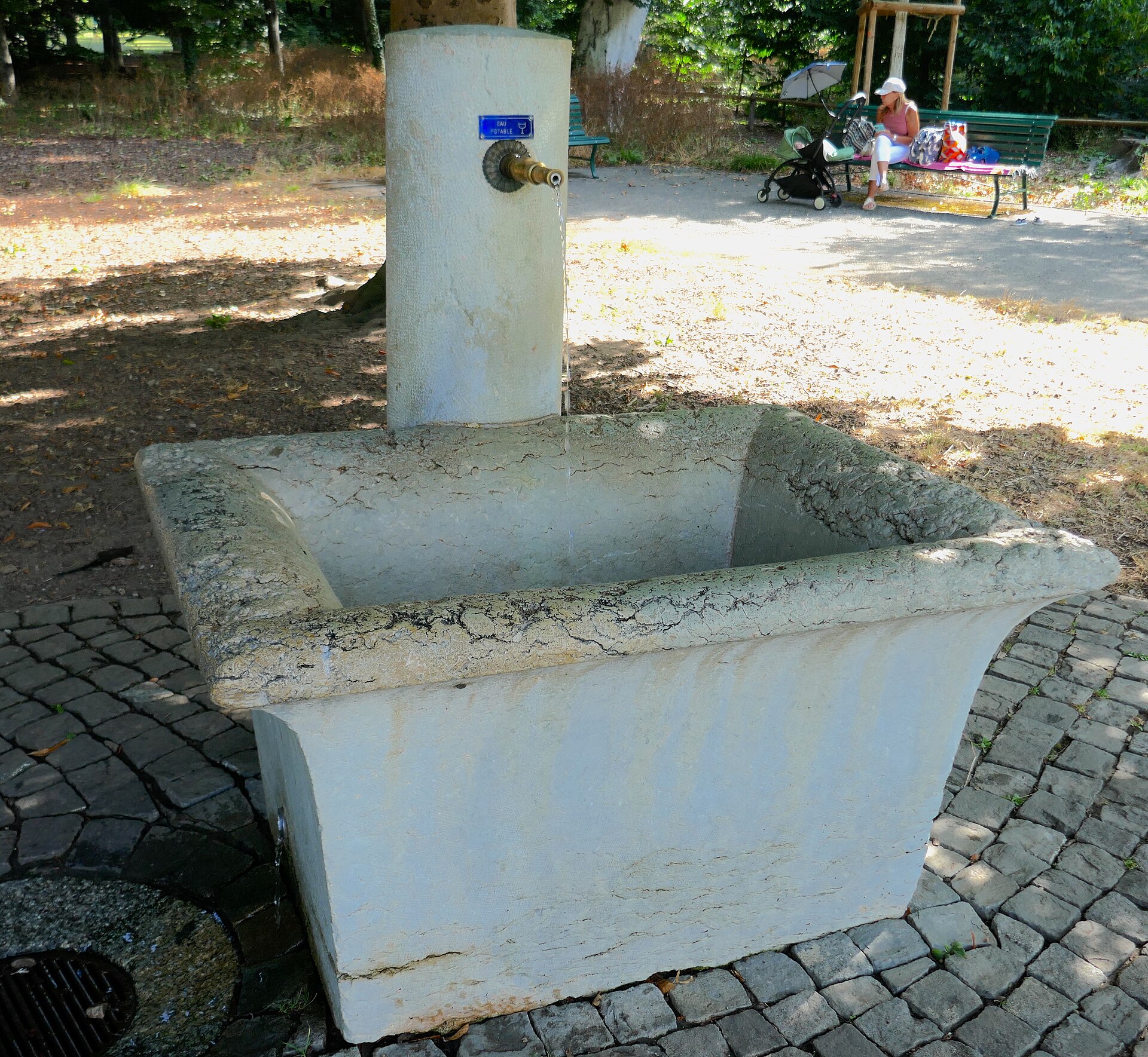 A white stone drinking fountain with a brass tap on a cobbled base in Parc La Grange in Geneva, with a park bench and trees behind