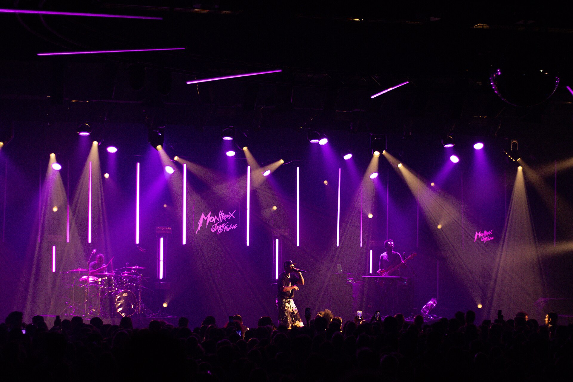 A performer on a stage lit with purple spotlights and vertical light bars at the Montreux Jazz Festival in 2026, with the audience in silhouette