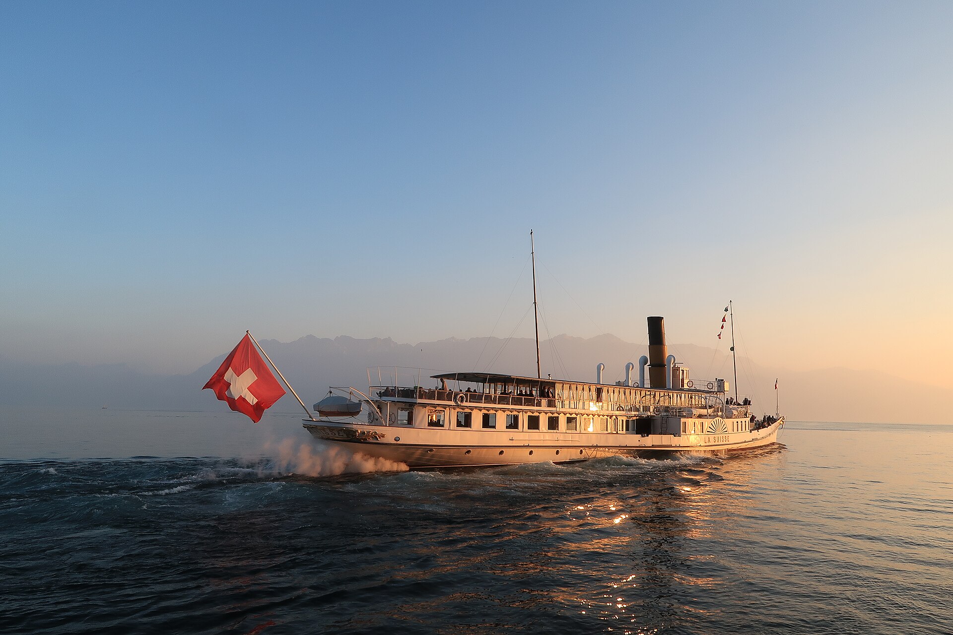 A Belle Époque paddle steamer leaving Cully in golden evening light, with a large Swiss flag at the stern and a glowing lake
