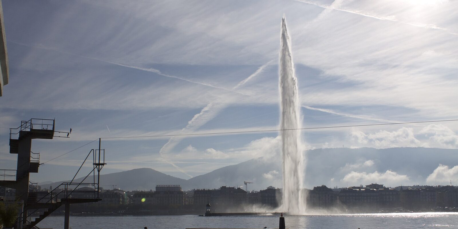 The Jet d'Eau rising against a streaked blue sky across Geneva's harbour, seen from the Bains des Pâquis with part of the diving tower on the left